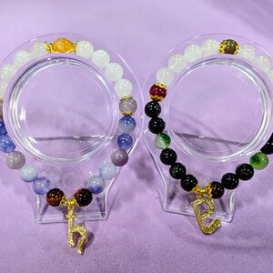 May include: Two beaded bracelets with charms. One bracelet has white and blue beads with a gold charm. The other bracelet has black, green, and red beads with a gold charm.