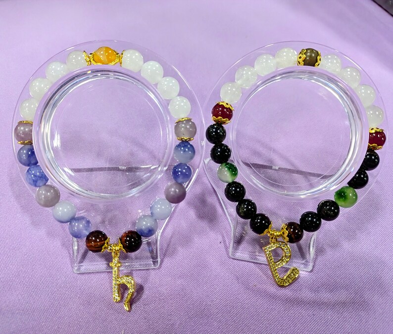 May include: Two beaded bracelets with charms. One bracelet has white and blue beads with a gold charm. The other bracelet has black, green, and red beads with a gold charm.
