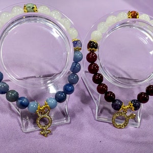 May include: Two bracelets with gemstone beads. One bracelet has blue and white beads with a gold charm. The other bracelet has red and white beads with a gold charm.