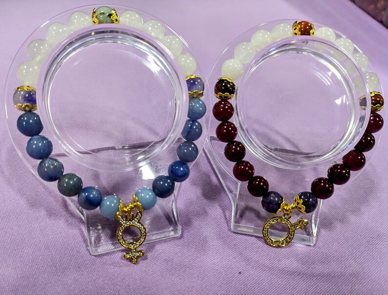 May include: Two bracelets with gemstone beads. One bracelet has blue and white beads with a gold charm. The other bracelet has red and white beads with a gold charm.