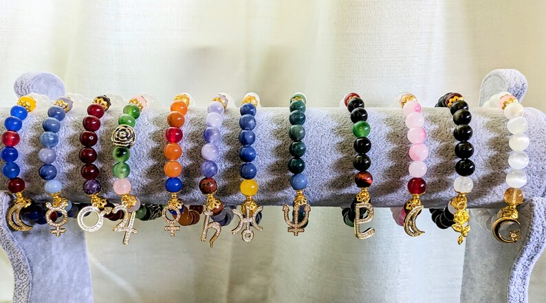 May include: A display of 12 colorful beaded bracelets with gold charms. Each bracelet features a different gemstone and a unique symbol, including a crescent moon, a star, a flower, and a trident.
