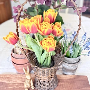 May include: A woven basket filled with vibrant orange and yellow tulips, with a decorative gold bunny figurine on a wooden board. Other potted flowers and greenery are also present, creating a spring-themed display.