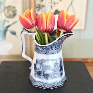 May include: A vintage blue and white ceramic pitcher with a landscape design, holding three tulips with red and yellow petals. The pitcher has a handle and is placed on a dark surface.