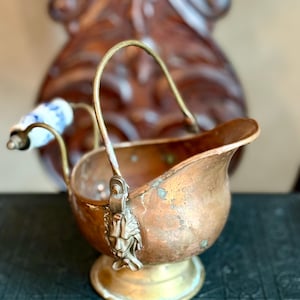 May include: Antique copper sugar bowl with a brass base and handle. The bowl features a lion's head detail and a blue and white porcelain handle. The bowl is a vintage item.