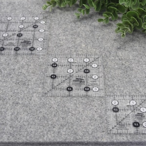 May include: Three clear acrylic quilting rulers with black and white markings are arranged on a gray felt surface. The rulers are marked with inch measurements and angles. Green foliage is in the upper right corner.