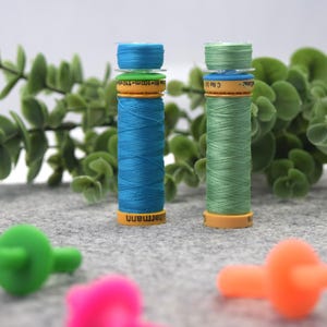 May include: Two spools of thread, one blue and one green, are displayed on a gray surface. The spools are labeled " Gütermann ".