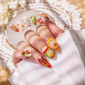 May include: Close-up of a hand with several fingernails painted with different food designs. The nails feature a red and yellow hot dog, a green salad with tomatoes, a white egg with a green salad, a red and white striped nail with a white egg, and a yellow pastry with a red tomato.