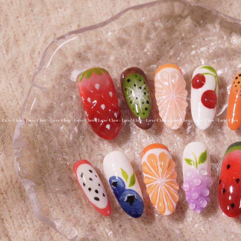 May include: Ten different nail designs featuring various fruits, including a strawberry, kiwi, orange, cherry, grapes, watermelon, and a dragon fruit. The nails are painted on a white background.
