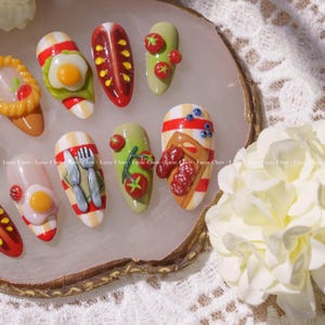 May include: A set of ten false fingernails decorated with miniature food items, including eggs, bacon, tomatoes, and a slice of bread with sausage. The nails are arranged on a white and gold tray.