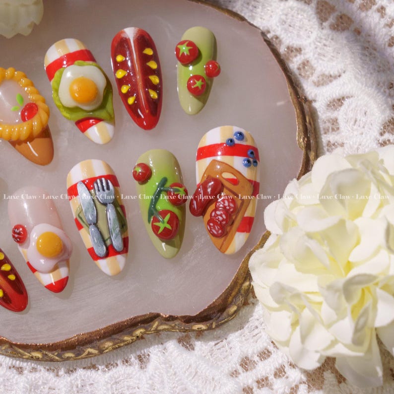May include: A set of ten false fingernails decorated with miniature food items, including eggs, bacon, tomatoes, and a slice of bread with sausage. The nails are arranged on a white and gold tray.