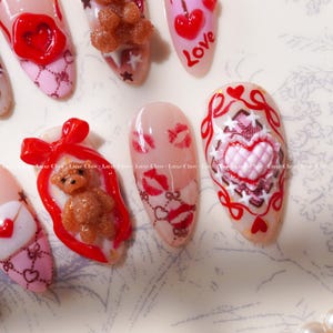 Peut inclure: Une collection d'ongles artificiels avec divers motifs. Certains ongles pr&eacute;sentent des c&oelig;urs rouges, des ours en peluche et le mot "Love". D'autres affichent des marques de baisers et des motifs d&eacute;coratifs roses et rouges.