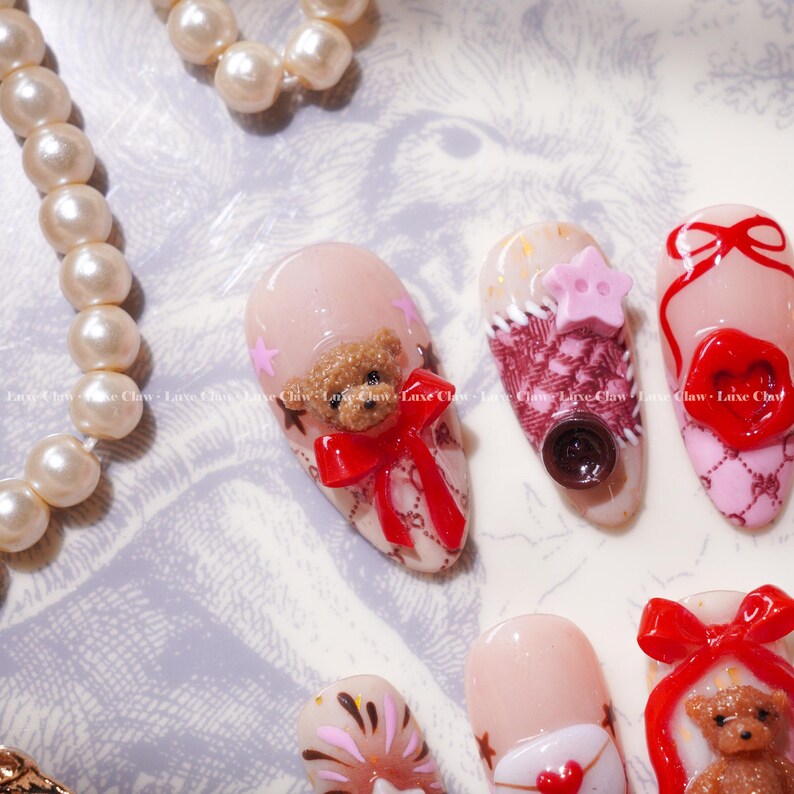 Peut inclure: Gros plan sur plusieurs ongles manucur&eacute;s avec divers motifs. Un ongle pr&eacute;sente un petit ours en peluche avec un n&oelig;ud rouge. D'autres ongles ont des &eacute;toiles roses, un bouton et un motif en forme de c&oelig;ur. Un collier de perles est en arri&egrave;re-plan.
