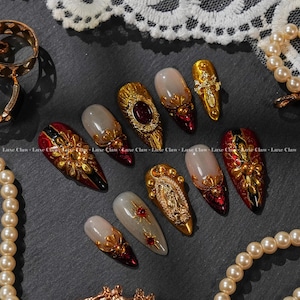 May include: An assortment of elaborately decorated artificial nails. The nails are long and feature gold detailing, including floral patterns, religious imagery, and jewel accents. They have a glossy finish with red, black, and nude tones.