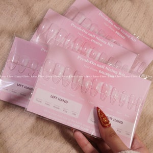 Op de afbeelding: Een roze en witte nagelmaatset met doorzichtige kunstnageltips. De set bevat drie verschillende nagelvormen: vierkant, stiletto en ballerina. De set bevat ook een kaart om de nagelmaten te meten.