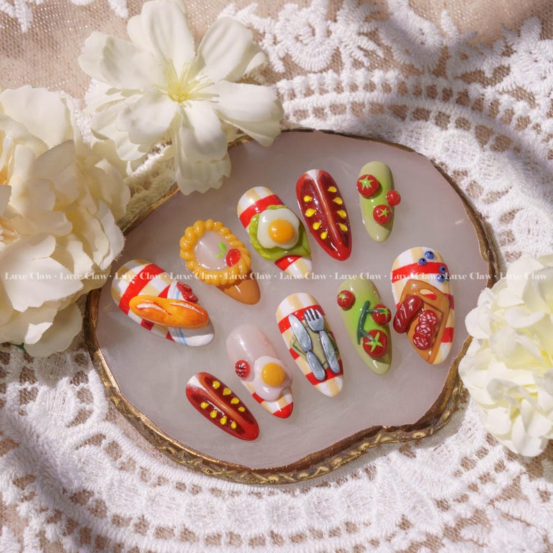 May include: A set of ten fake nails with different food designs. The nails are painted with red and white stripes, and each nail has a different food design, including a fried egg, a hot dog, a plate of fries, a sandwich, and a plate of cutlery.