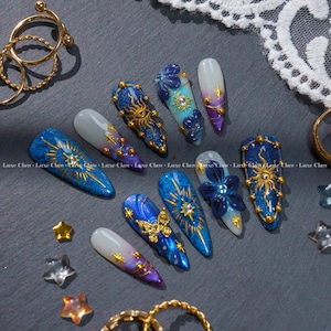 Peut inclure: Une collection de faux ongles longs en forme d'amande avec des motifs complexes. Les ongles présentent des thèmes célestes avec des soleils dorés, des étoiles et des papillons sur un fond dégradé bleu, violet et blanc. Certains ongles ont des ornements floraux en 3D.