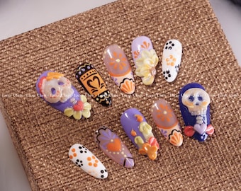 May include: Ten purple and white false fingernails with various designs, including skulls, flowers, and hearts. The nails are arranged on a brown burlap surface.
