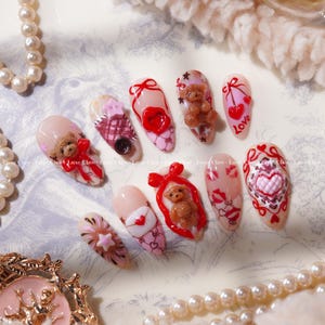 Peut inclure: Un ensemble de dix faux ongles avec divers motifs. Les ongles pr&eacute;sentent des ours en peluche, des c&oelig;urs, des n&oelig;uds et le mot "Love". La palette de couleurs comprend le rose, le rouge et le marron, certains ongles ayant une finition paillet&eacute;e. Les ongles sont dispos&eacute;s sur une surface d&eacute;corative.