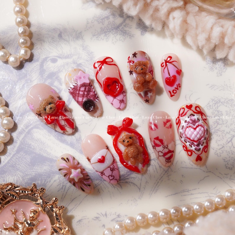Peut inclure: Un ensemble de dix faux ongles avec divers motifs. Les ongles pr&eacute;sentent des ours en peluche, des c&oelig;urs, des n&oelig;uds et le mot "Love". La palette de couleurs comprend le rose, le rouge et le marron, certains ongles ayant une finition paillet&eacute;e. Les ongles sont dispos&eacute;s sur une surface d&eacute;corative.
