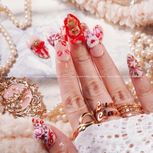 Peut inclure: Une main avec de longs ongles d&eacute;cor&eacute;s. Les ongles pr&eacute;sentent divers motifs, notamment des ours en peluche, des c&oelig;urs et des empreintes de l&egrave;vres. Les ongles sont peints dans des tons de rose, de rouge et de blanc, avec des bagues dor&eacute;es aux doigts. Le texte "Luxe Claw" est visible.