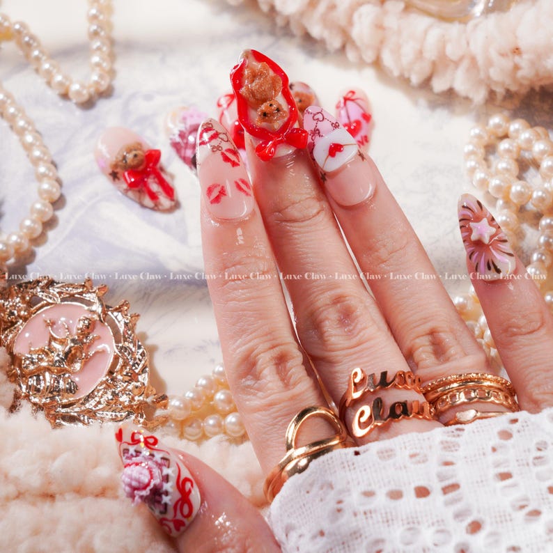 Peut inclure: Une main avec de longs ongles d&eacute;cor&eacute;s. Les ongles pr&eacute;sentent divers motifs, notamment des ours en peluche, des c&oelig;urs et des empreintes de l&egrave;vres. Les ongles sont peints dans des tons de rose, de rouge et de blanc, avec des bagues dor&eacute;es aux doigts. Le texte "Luxe Claw" est visible.