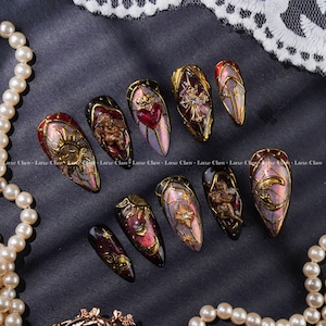 Puede incluir: Un conjunto de diez uñas postizas largas en forma de almendra con diseños intrincados. Cada uña presenta un borde dorado y un diseño único, que incluye soles, lunas, corazones, cruces y querubines. Las uñas tienen una combinación de colores rojo oscuro, dorado y morado.
