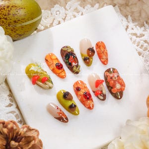 May include: Ten artificial nails with autumnal designs. The nails showcase detailed artwork including acorns, ladybirds, mushrooms, a fox, and a teddy bear. Colours include orange, brown, yellow, and beige, with intricate detailing.