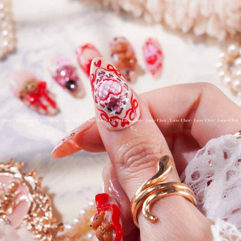 Peut inclure: Une main avec de longs ongles en amande, chacun orn&eacute; d'un nail art complexe. Les motifs comprennent des ours en peluche, des c&oelig;urs et des motifs floraux dans des tons de rouge, de rose et de marron. Une bague serpent dor&eacute;e est sur le doigt. Les ongles sont d&eacute;cor&eacute;s du texte "Luxe Claw".