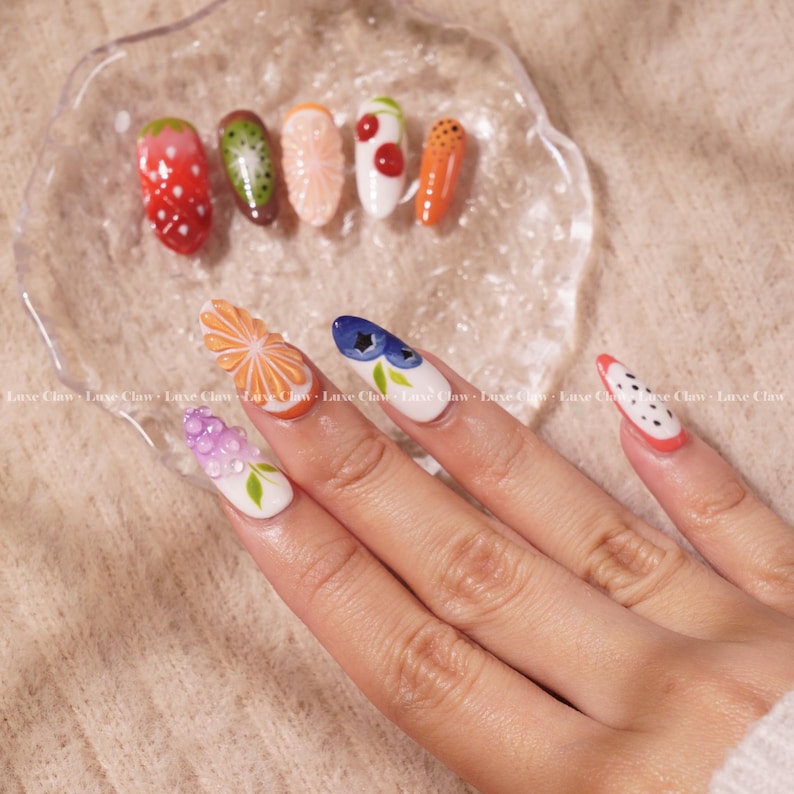 May include: A hand with long, almond-shaped nails painted with a variety of fruit and flower designs. The nails are painted with a white base and feature designs of strawberries, kiwi, oranges, cherries, carrots, flowers, blueberries, and polka dots.