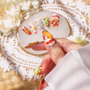 May include: Close-up of a hand with painted nails featuring a variety of food designs. The nails are painted with images of hot dogs, french fries, eggs, and other food items. The nails are on a white background.