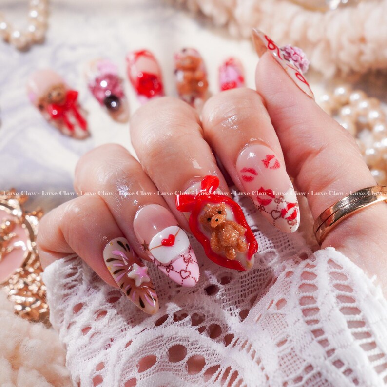 Peut inclure: Une manucure avec plusieurs ongles en acrylique aux motifs vari&eacute;s. Certains ongles pr&eacute;sentent un ours en peluche dans un cadeau rouge, tandis que d'autres arborent des motifs de c&oelig;urs et de baisers. Les ongles sont peints en rose, rouge et marron, certains orn&eacute;s de n&oelig;uds.