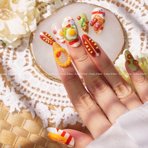 May include: A hand with colorful nail art featuring miniature food designs. The nails are decorated with designs of a pizza, a hot dog, a breakfast sandwich, a pie, and a tomato.