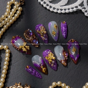 Pode incluir: Uma coleção de unhas postiças personalizadas em várias formas e tamanhos. As unhas apresentam um esquema de cores roxo e dourado com designs intrincados, incluindo enfeites dourados, pérolas e detalhes de estrelas. O texto "Luxe Claw" é visível.