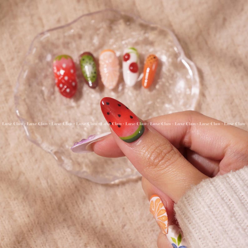 May include: Close-up of a hand with painted nails featuring various fruit designs. The nails are painted with watermelon, kiwi, orange, strawberry, and carrot designs.