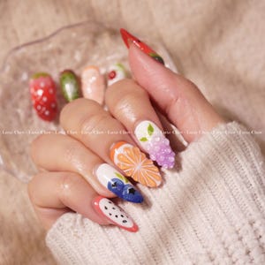 May include: Close-up of a hand with long, almond-shaped nails painted with a fruit-themed design. The nails are decorated with 3D fruit designs, including strawberries, watermelon, blueberries, grapes, and an orange slice.