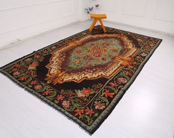 May include: A vintage rug with a dark brown background and a central medallion featuring a floral design in shades of green, red, and yellow. The rug has a fringed border.