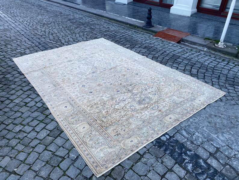May include: A large, off-white rug with a floral pattern and a border. The rug is laid out on a cobbled surface.