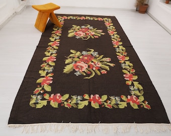 May include: A dark brown rug with a floral pattern. The rug has a white fringe border and features a repeating design of red, pink, and yellow flowers.