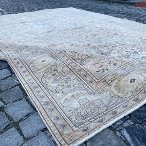 May include: A beige and cream coloured rug with a floral pattern and a border. The rug is lying on a cobbled street.