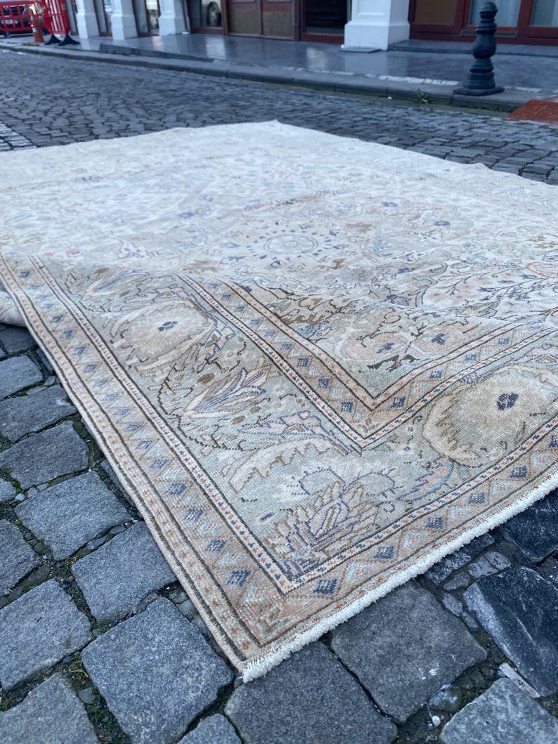 May include: A beige and cream coloured rug with a floral pattern and a border. The rug is lying on a cobbled street.