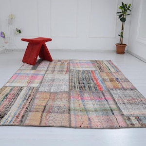 Puede incluir: Una alfombra de patchwork multicolor con una variedad de texturas y patrones. La alfombra está hecha de diferentes piezas de tela cosidas juntas. La alfombra está colocada sobre un suelo blanco con un taburete rojo en el fondo.