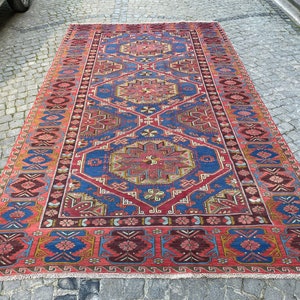 May include: A vintage Turkish rug with a red background and intricate blue, yellow, and brown geometric patterns. The rug has a fringed edge and is in good condition.