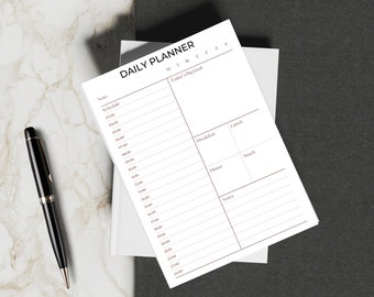 May include: A white daily planner with a black header that says "DAILY PLANNER". The planner has columns for each day of the week, a section for "Today's Big Goal", and sections for "Breakfast", "Lunch", "Dinner", and "Snack".