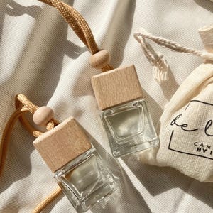 Könnte beinhalten: Zwei transparente Auto-Lufterfrischer aus Glas mit Holzdeckeln und beigefarbenen Kordeln. Ein kleiner beiger Kordelzugbeutel mit schwarzem Text ist ebenfalls im Bild. Die Lufterfrischer sind quadratisch und scheinen leer zu sein.