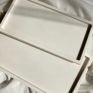 Könnte beinhalten: Zwei weiße rechteckige Tabletts, die übereinander gestapelt sind. Die Tabletts bestehen aus einem glatten, leicht strukturierten Material.