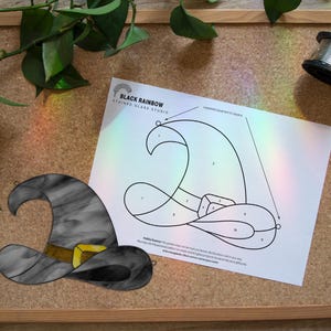 Op de afbeelding: Een gebrandschilderde heksenhoed met een gouden gesp, naast een patroon om een ​​vergelijkbare hoed te maken. Het patroon staat op een wit vel papier. Een spoel draad ligt op een prikbord, met groene bladeren op de achtergrond.