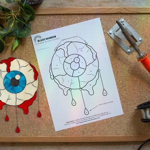 Op de afbeelding: Een glas-in-oog met een blauwe iris en rode accenten, naast een patroon en een soldeerbout. Het oogontwerp staat op een houten frame, met de tekst "Black Rainbow Stained Glass Studio".