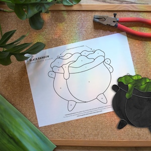 Può includere: Un progetto artigianale con un calderone nero con accenti verdi gocciolanti, accanto a un disegno a tratto dello stesso calderone. Una lastra di vetro verde, una pinza e una pianta sono presenti su una bacheca di sughero.