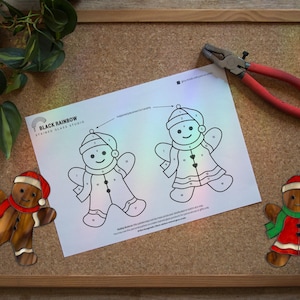 Op de afbeelding: Een prikbord met een houten frame toont een ontwerp van "Black Rainbow Stained Glass Studio", een tekening van een peperkoeken man en een tang. Er zijn ook twee peperkoeken man ornamenten aanwezig, een met een rode sjaal en de andere met een groene sjaal.