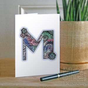 Könnte beinhalten: Weiße Grußkarte mit einem verzierten Buchstaben "M" in einem Paisley-Muster in Rosa, Blau und Grün. Ein schwarzer Stift liegt auf einer Holzoberfläche. Im Hintergrund befindet sich ein dekorativer Topf mit einer Pflanze. Die Karte ist von Melodel Designs.